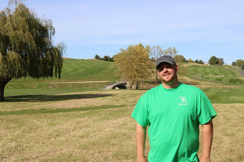 Andy Young wearing a gray Nike hat and a green Woodbine Bend shirt smiles with the Woodbine Bend golf course as his backdrop