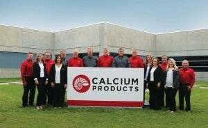 Fifteen men and women in black, gray, red, and white outfits smile while standing behind a large red and white Calcium Products sign in front of the concrete Calcium Products building