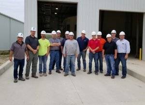 Thirteen men wearing safety glasses and white hard hats stand in front of a gray warehouse and smile for a photo