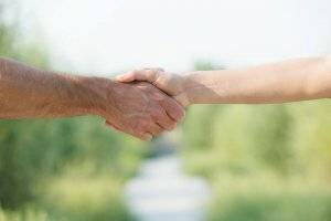 The forearm and hand of an older man shaking hands with the forearm and hand of a younger man in the middle of a field