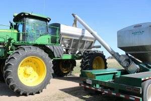 A green and silver Doyle truck is dispensing fertilizer into the back of a green John Deere tractor so it can go out into the field